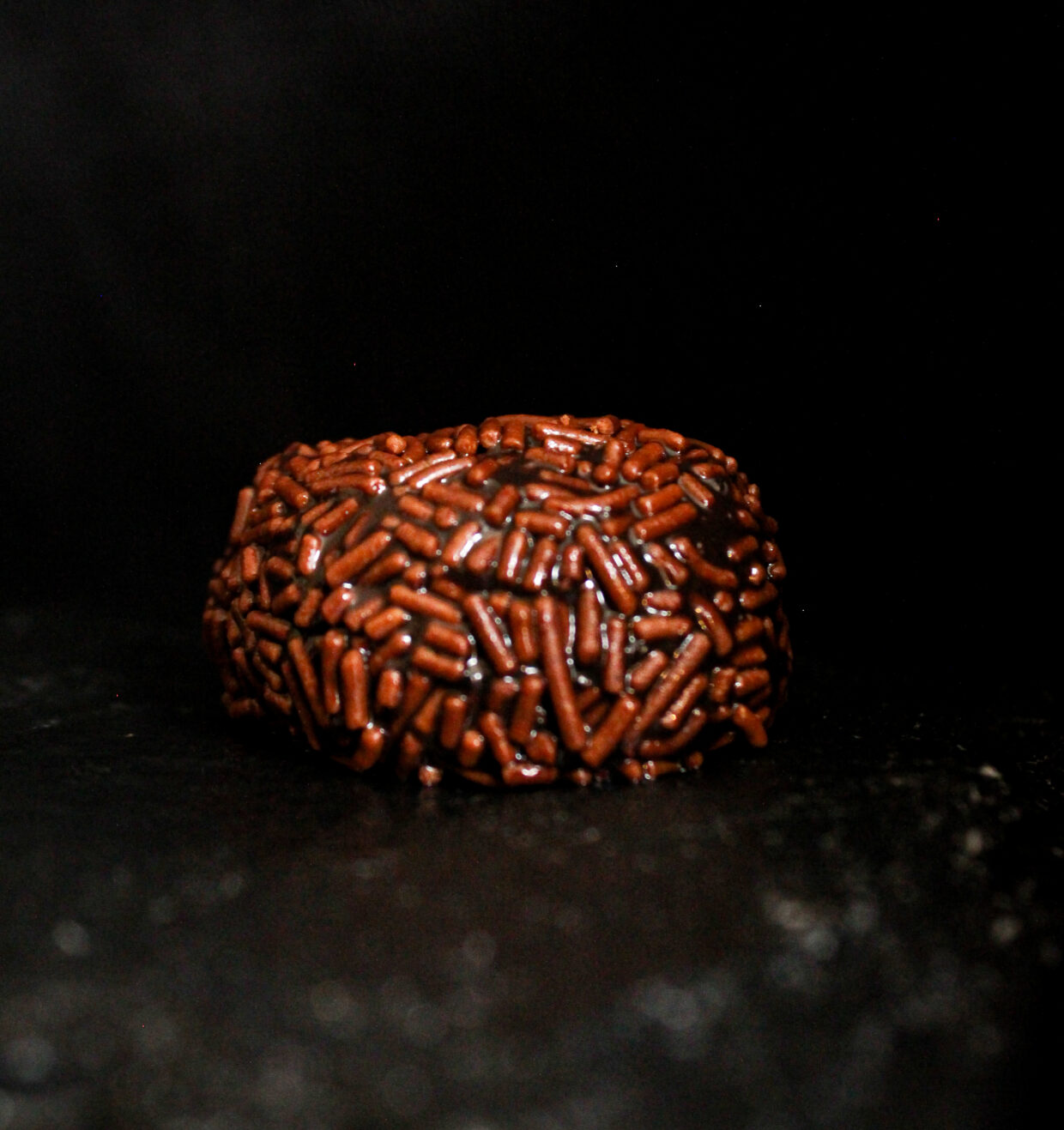 image description; The photo is taken on a dark background, we can see one truffle sat on a piece of slate, the truffle is rich dark brown in colour and is covered in chocolate vermicelli sprinkles which are a little lighter in colour, the truffle is reflecting the light a