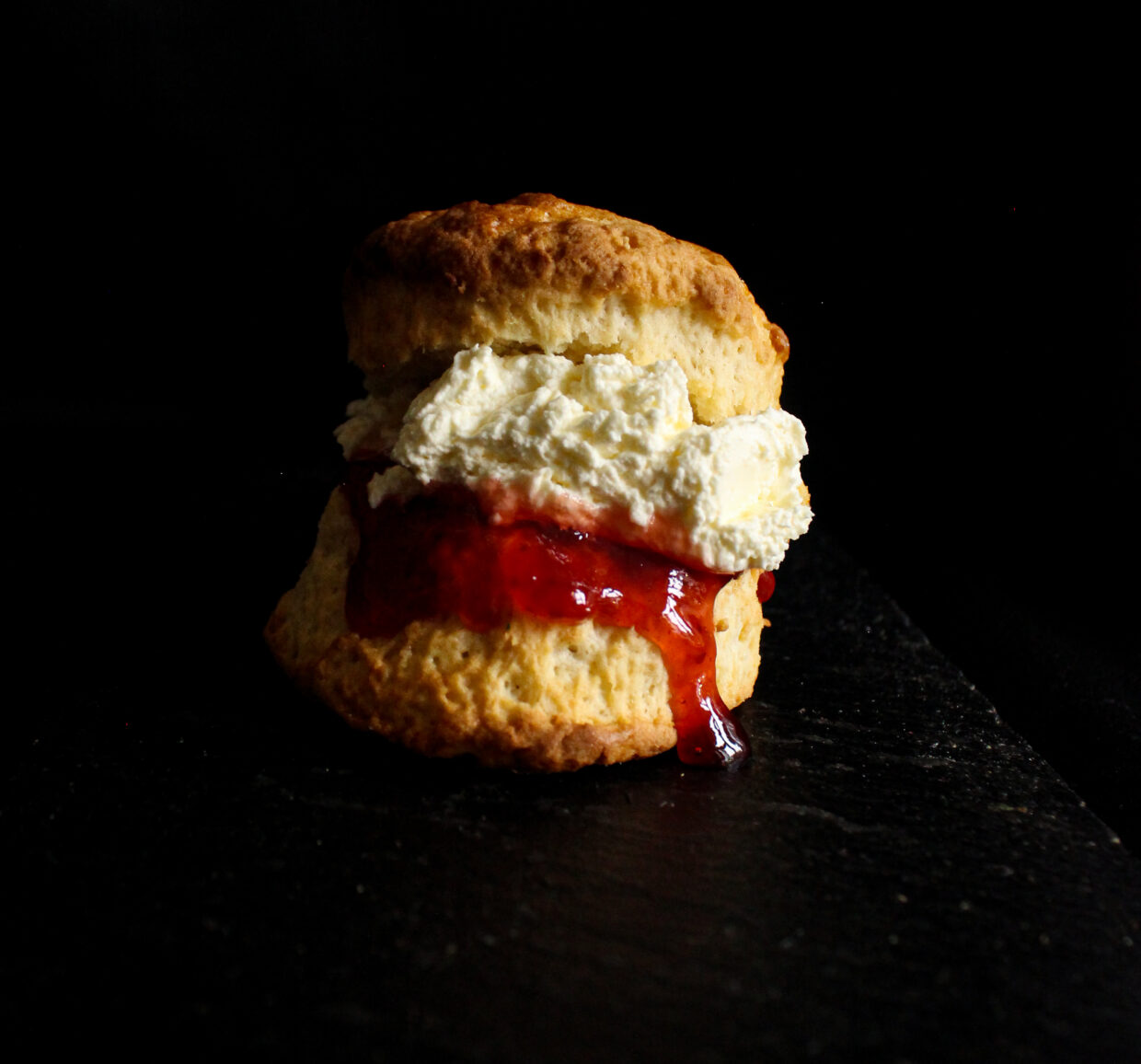 image description: on a dark background we can see a scone, it is filled with deep red strawberry jam and cream. The jam is dripping down one side of the scone and the cream is on top, the scone is golden brown and quite tall.