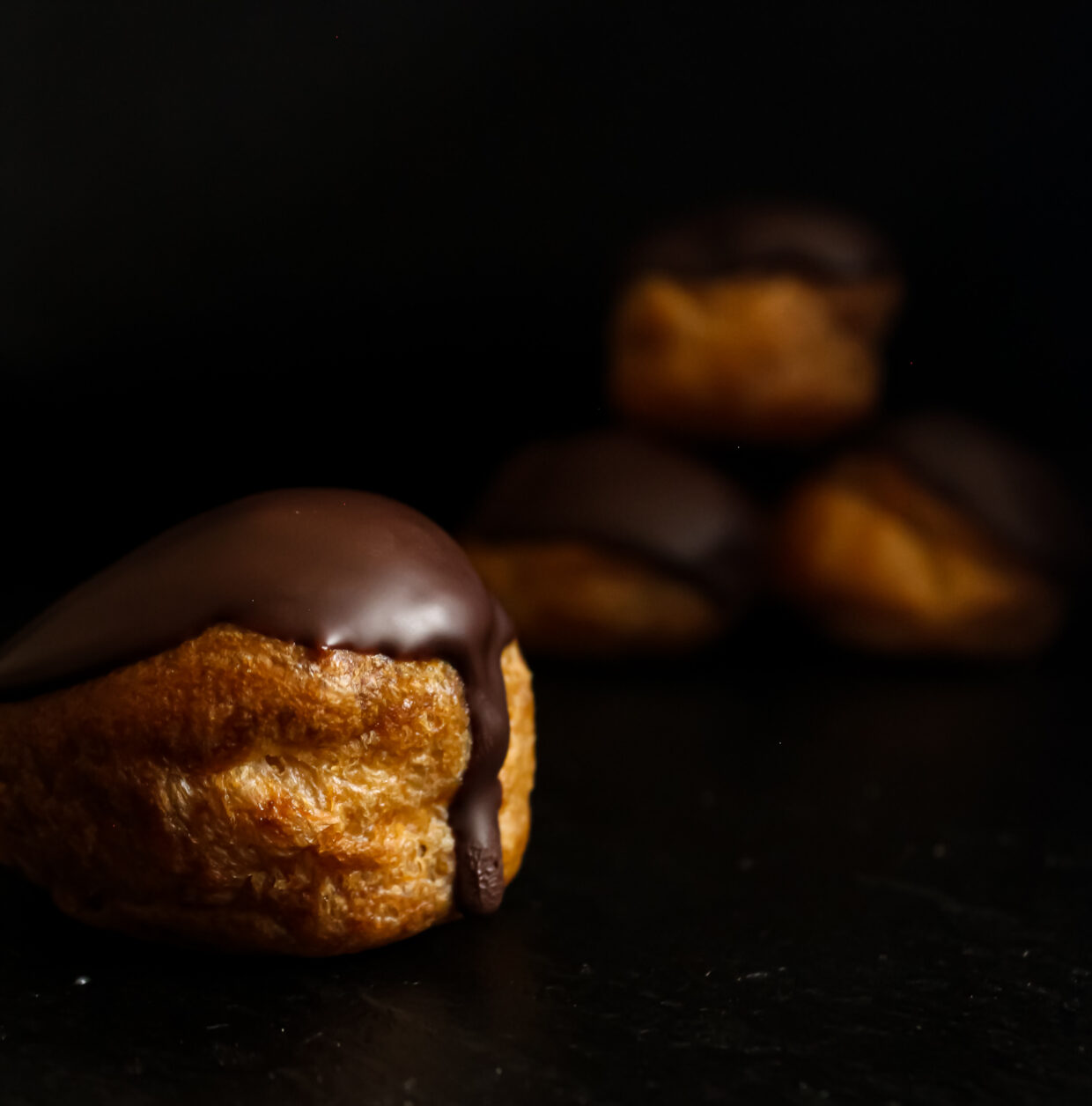 image description: Set on a dark background, we can see one profiterole in the foreground and three stacked in the background, blurred out. The Profiterole in the foreground has a chocolate dipped top, the chocolate has now set but there is one drip coming down the front of the pastry and we can see condensation where it has just come out of the fridge. The light is reflecting of the chocolate and the pastry is a nutty but light golden colour.