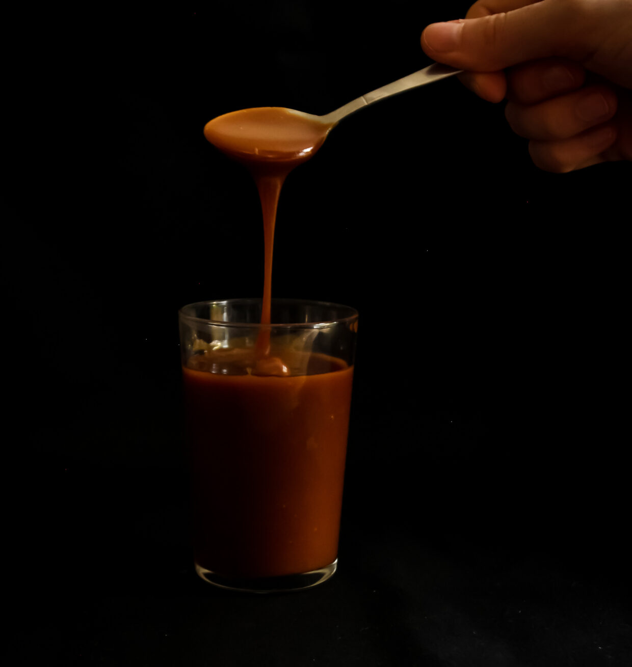 image description; Set on a black background we can see a glass filled with golden brown salted caramel, above the glass there is a spoon and ribbons of salted caramel are falling back into the glass reflecting the light.
