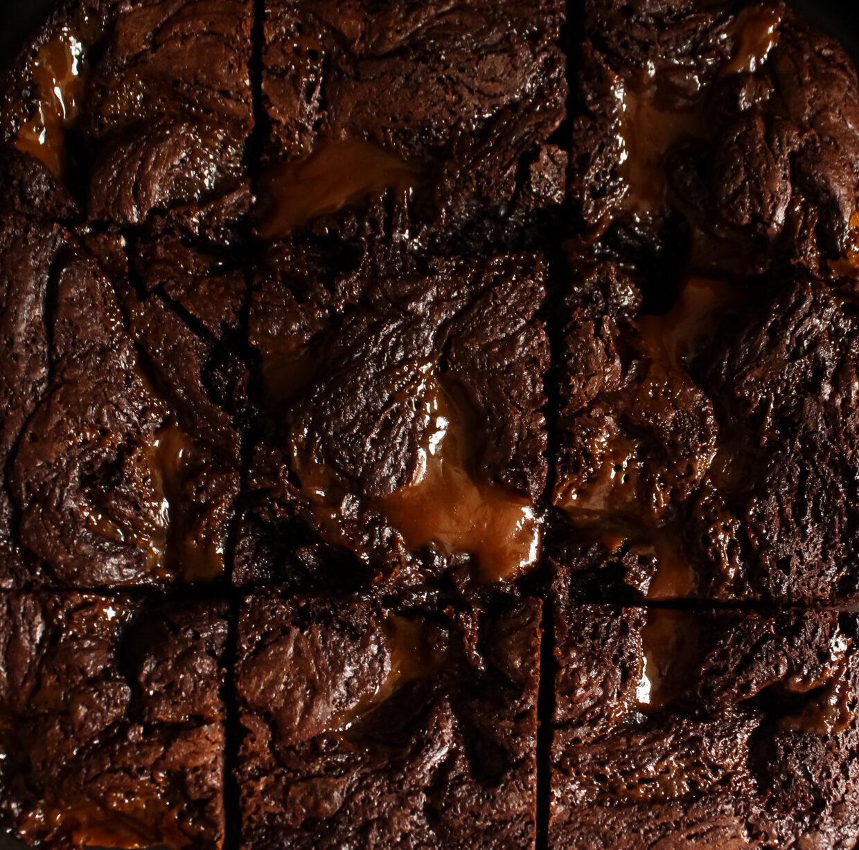 image description: the shot is taken from over head and the brownies fill the screen, as if freshly sliced there are nine brownies pushed closely together, the brownies have a slightly cracked and textured top giving them a rustic appearance, they are dark rich brown in colour and have light salted caramel swirled into the batter glistening and reflecting the light slightly.